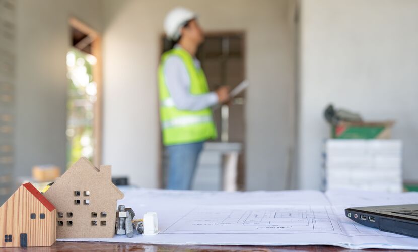 Maquette en bois de maison, vis et plans de construction sur une table, avec un ingénieur en arrière-plan observant des plans dans un chantier.